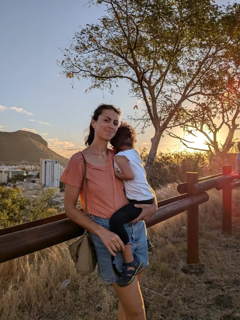 Célia Duwicquet tenant sa fille dans ses bras au coucher du soleil, sur un sentier avec vue sur la ville et la montagne à l’arrière-plan.